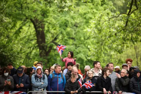 Reuters A small girl wearing a pink jumper waves a Union Jack in her hand while on the shoulders of a member of the crowd lining the Mall, with other members of the public around them and green foliage in the background.