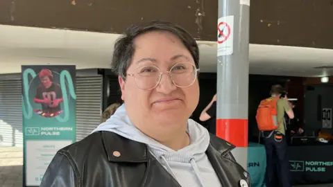 Kay Davidson/BBC Jo Watson, a woman with short black hair and round glasses, looks at the camera. She is slightly smiling. She is wearing a grey hoodie and a black leather jacket. There is a grey pole to the left of her and a light blue banner which says Northern Pulse to her right.