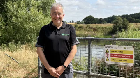A man with white hair is smiling. He is wearing a black t-shirt with a green logo that reads "environment agency". He is standing near metal fencing with warning signs on it. Beyond him is the river Severn surrounded by green and brown grass