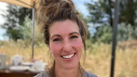 Alice Kennish - a woman smiling at the camera. She has a mass of light brown hair tied up on her head.