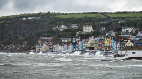 Getty Images A stormy sea batters a prominate along which is a row of colourful houses. In the background some green fields and above a cloudy sky