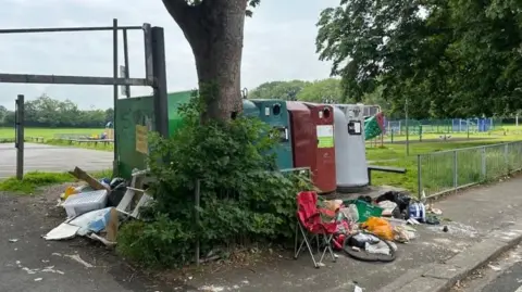 A row of large recycling bins in a public car park with an assortment of items, including plastic bags, boxes and a camping chair, dumped around them.