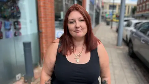 Becki Bowden/BBC A woman with red hair and a black vest stands on a street next to parked cars. Shop fronts are also visible on the left of the image.