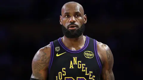 LeBron James stands in front of a black background. He is bald, has a black beard and has heavily tattooed arms. He is wearing a black basketball top which says Los Angeles in yellow letters.