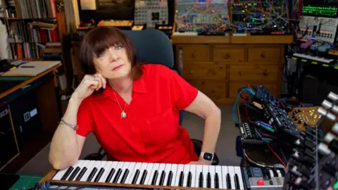Chris Carter Cosey Fanni Tutti sitting in front of a keyboard. Behind her are racks of music related electronics. She is in a home studio. She is wearing a red shirt.