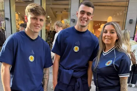 Rachael Vaughan Three people pose in Scotland tops. Left is Ben Gannon-Doak who has short brown hair and tattoos on one arm and next to him is Lennon Miller who is slightly taller and has a navy jumper tied around his waist. On the far right is Rachael who has long dark blonde hair and tattoos on one arm