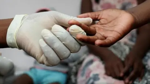 A glove-wearing nurse takes a blood sample from a child for an HIV test while the child's mother looks on at a clinic in Diepsloot, north of Johannesburg, South Africa, 12 March2025