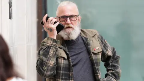 A man with long white hair, a full white beard and glasses holds a mobile phone to one ear while standing outside a building. The person is wearing a checked overshirt over a dark T-shirt, with tattoos visible on one hand.