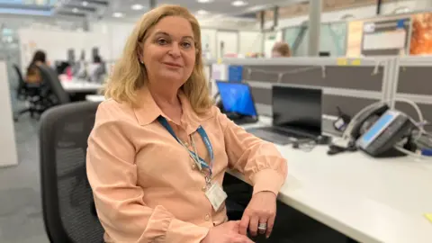 Seb Cheer/BBC Fiona McCulloch, the Chief Officer York Citizens Advice, is sitting in an office chair, leaning her left arm on a desk. There are people in the background working at computers.