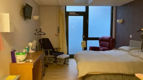 The inside of a hospital room with a double bed, a desk and two seats with purple covers.