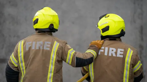 Two firefighters with their back to the camera. They are both wearing a high-vis yellow helmet and brown jackets with "fire" written on the back. One firefighter has their hand on the other's shoulder.