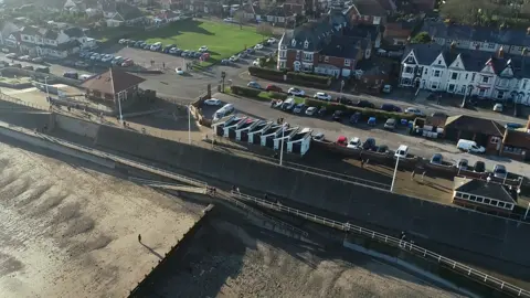 A drone image showing golden sands and a seafront dotted ith houses and huts.