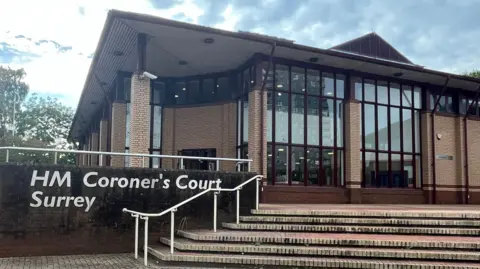 A general view of HM Coroner's Court Surrey. It is a brown brick building with outdoor stairs leading to the buildings facade which has long narrow windows. There are large white letters on a wall reading HM Coroner's Court Surrey.