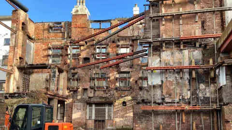 The remaining exterior walls and brickwork of an old fire ravaged building, with an orange and black digger in the foreground.