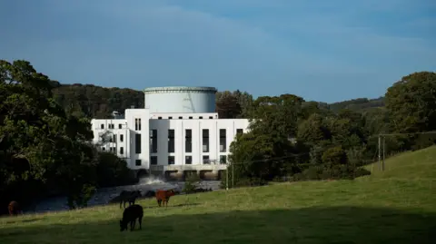 Drax A view of the white art deco Tongland hydro power station building nestling in among trees with cattle grazing in front of it