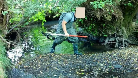 Natalie Barstow Picture taken from a video captured on a wildlife camera shows someone walking through a stream in waterproof trousers and carrying a net.  Their face is obscured.
