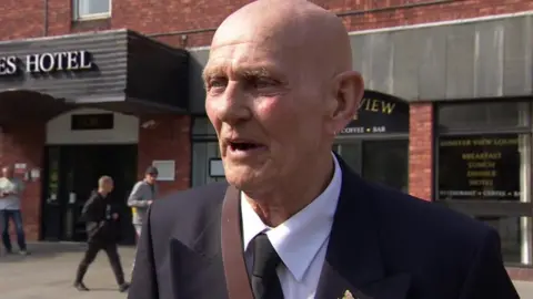 A man with a bald head wearing a blue suit jacket with a white shirt and black tie is talking to the BBC.