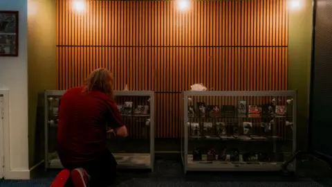 Simon Shaw Adam Parkin is crouching in front of one of two cabinets lined with photos. There is a wooden, slatted design on the wall behind him and three bare lightbulbs shining over head.