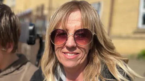 Janet is sitting down and is in the middle. She has long blonde hair and a fringe and is wearing sunglasses and a black coat.