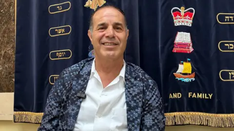 Michel Dadoun is seen smiling, wearing a blue patterned blazer over a white shirt, sitting in front of a dark blue velvet backdrop featuring gold Hebrew lettering and a crown emblem.