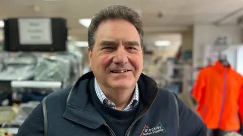 George Carden/BBC James MacGregor wearing a jumper with a shirt underneath smiling in his shop with shirts on shelves behind him. He has brown hair