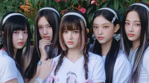 Promotional photo of the band NewJeans, with the five members dressed in white t-shirts, with white alice bands in their hair, against a floral backdrop.