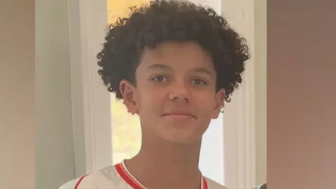 Noah smiling at the camera. He is wearing a white football shirt with red edging and has short curly hair.