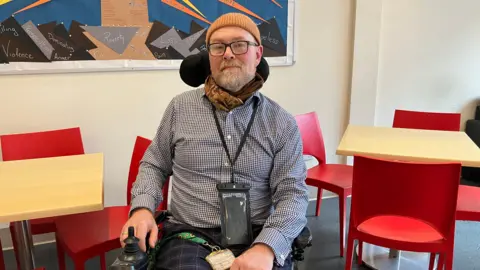 A middle-aged man wearing a black and white checked shirt and light brown beanie hat sitting in a wheelchair. He is inside in a room with small tables surrounded by chairs. On the wall there is a mood board with words written on it such as poverty, loss and violence.