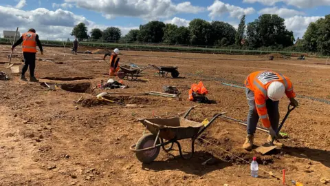 Mola A dig site on a levelled field. A small group of people work on digging with spades and are surrounded by wheelbarrows and holes in the earth.