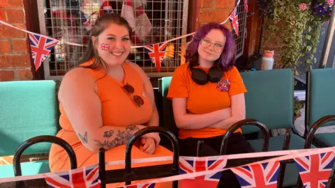 Two women are sitting outside on chairs. They are both wearing orange outfits and have Union Jack temporary tattoos on their faces.