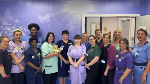 Northamptonshire Health Charity Thirteen women standing against a blue wall with a dandelion clock and butterflies on it and behind them a door. They are mostly in NHS uniforms, in shades of blue, lilac, light green and purple. They are looking towards the viewer and smiling. 