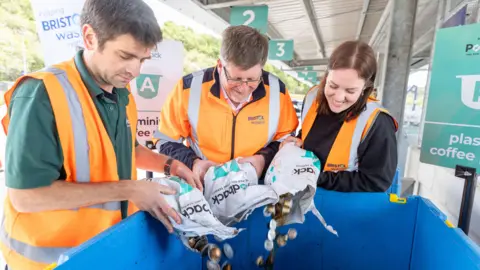 Barbara Evripidou Three people wearing orange high-vis jackets pouring used coffee pods into special waste bins.