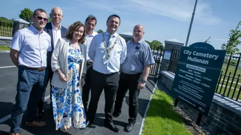 Six people, including the mayor Ruairí McHugh and a number of council officials, are standing beside the cemetery sign that reads: City Cemetery Mullenan. Black gates and a grassy embankment are visible in the background. 