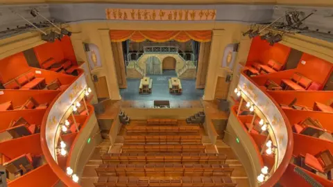 David P Howard/Geograph An aerial view of the inside of the Theatre Royal in Bury St Edmunds. There is red seating in the gallery and the wooden floorboards of the stage can be seen.