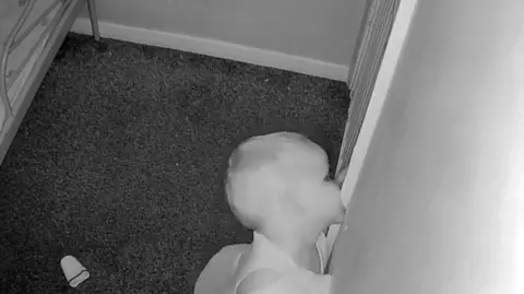 Jess Harry Una imagen fija en blanco y negro de un bebé montiro de un niño mordiendo el marco de una puerta blanca. Detrás de él está su armazón de cama de metal.