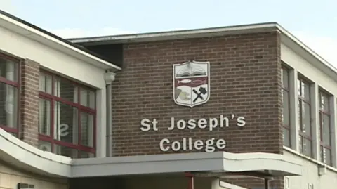 A large school crest is hung on the side of an old red-bricked building. Underneath it is a sign which reads 'St Joseph's College' in white text. The crest depicts a book, the outline of two fish facing each other, a hand reaching out open palmed and a hammer and chisel crossed over each other.