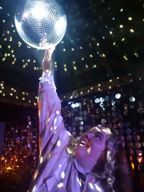 Duncan Mitchell A young girl holds her hand up to a disco ball surrounded by coloured lights.