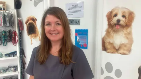 A woman with long brown hair smiles at the camera. She is wearing a grey dress. Behind her is a white shelf with dog collars hanging on it, and two pictures of dogs with paw print stickers on a wall behind her.