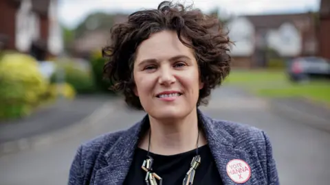 PA Claire has a blurry background behind her of a housing development. 

She has wiry short brown hair and is wearing red lipstick, grey blazer and black top. 
She is wearing a chunky metallic necklace. 