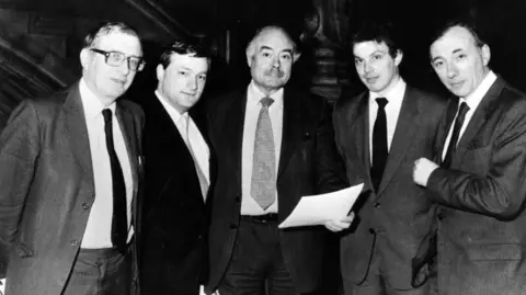 Keystone/Getty Images A black and white photo shows five men standing side-by-side. They are all wearing suits and ties and the man in the centre is holding a sheet of paper.