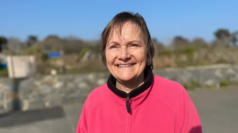 Val Goodwin - A lady with a pink zip up jumper on, with a black rim. She has short black hair, and is smiling at the camera.