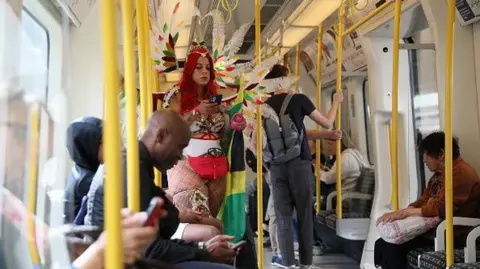 Getty Images A woman with long read hair and wearing a feathered head dress, is looking at her phone while stood on a London Underground train. Other Tube passengers, not dressed in Carnival clothes, are seated around her.