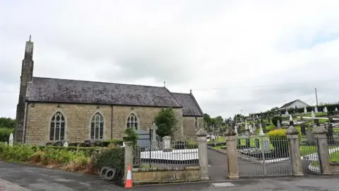 Pacemaker St Mary's Church in Maguiresbridge, with graveyard beside it containing grave stones. There are grey gates in front, with an orange and white traffic cone to the left.