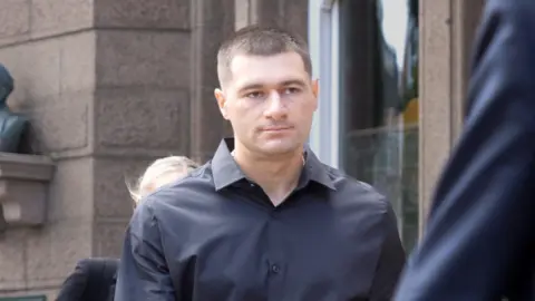Artur Sevash-Zade wearing a black shirt walks into court. He has short black hair and is clean shaven.