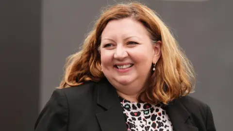 PA Media A woman with red and blonde hair, smiling while walking. She is wearing a dark jacket and a patterned top with various colours.