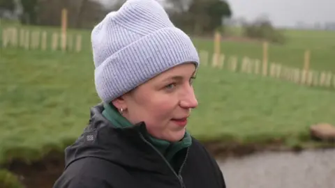 Hannah Lederer standing in a field by the pond. She is wealing a lilac knitted hat, green fleece and black coat