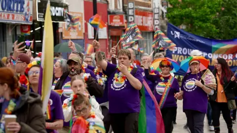 A gathering of people wearing purple and other brightly coloured clothing, walking in the street with rainbow flags
