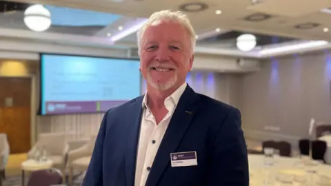 Lee Madden, a man with short white hair and a white, goatee-style beard. He is wearing a blue blazer over a white shirt with blue buttons. He is standing in a conference room, in front of a large projector screen. He is looking directly at the camera and smiling.
