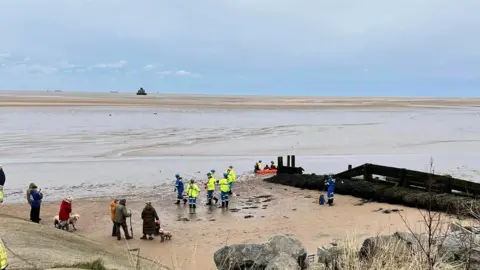 Julie Favell Connell The Humberston Fitties beach with bystanders watching on the sand as a team of emergency workers rescue a man from the mud. A wide expanse of water and sand is visible in the distance as is the Humber estuary which has a number of ships sailing on it.