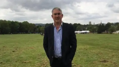 Scott Weston standing in a field wearing a suit jacket and striped blue shirt.